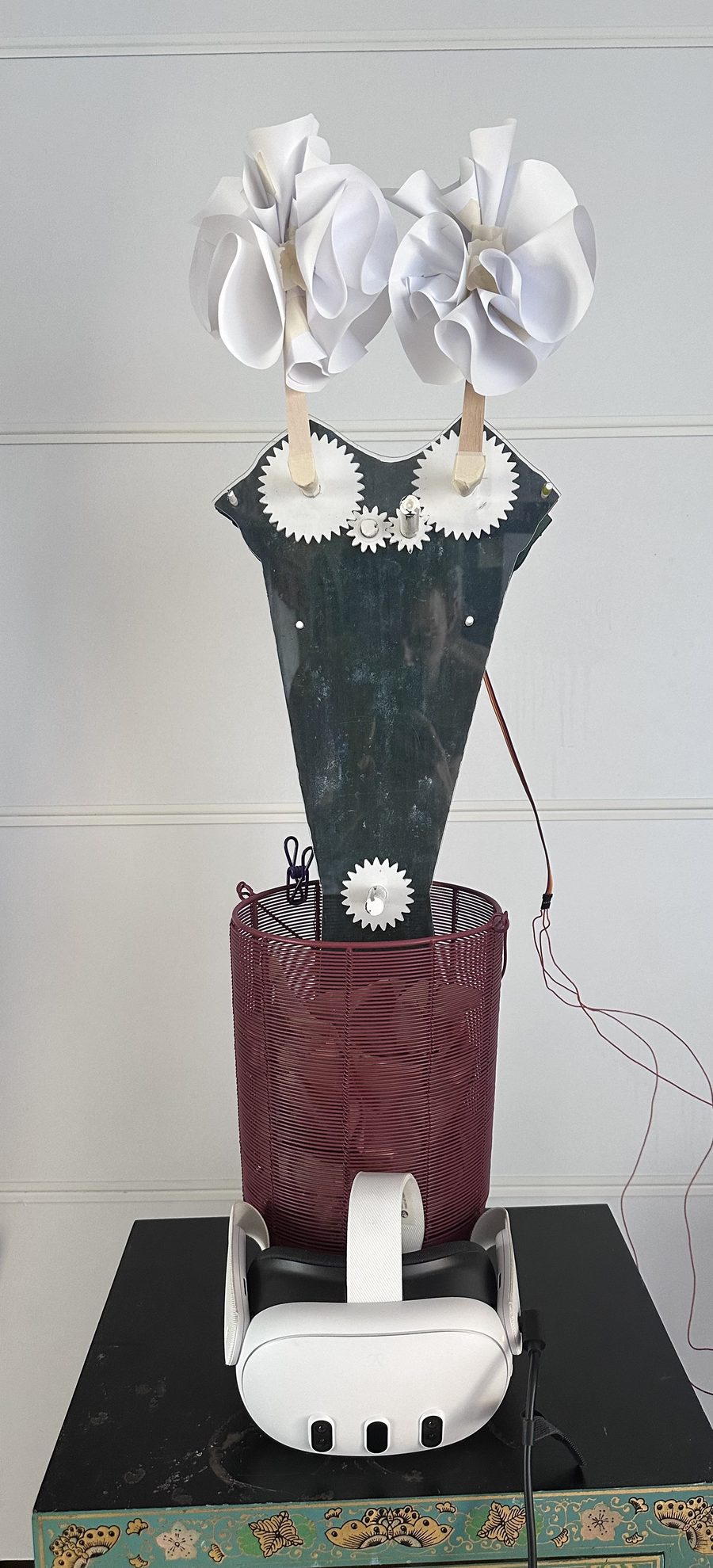 The re-posed sculpture: two paper flowers on gear-driven arms, a dark acrylic body in a red wire basket, a Quest 3 headset on the plinth below.