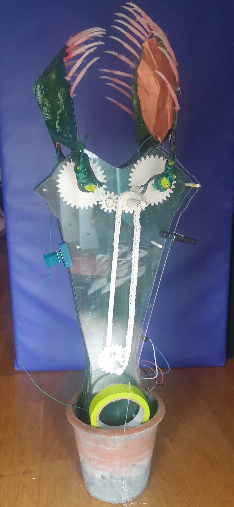 The first version of the sculpture: a clear acrylic body with white gears and a chain drive, green petals and a copper pad, standing in a clay pot.